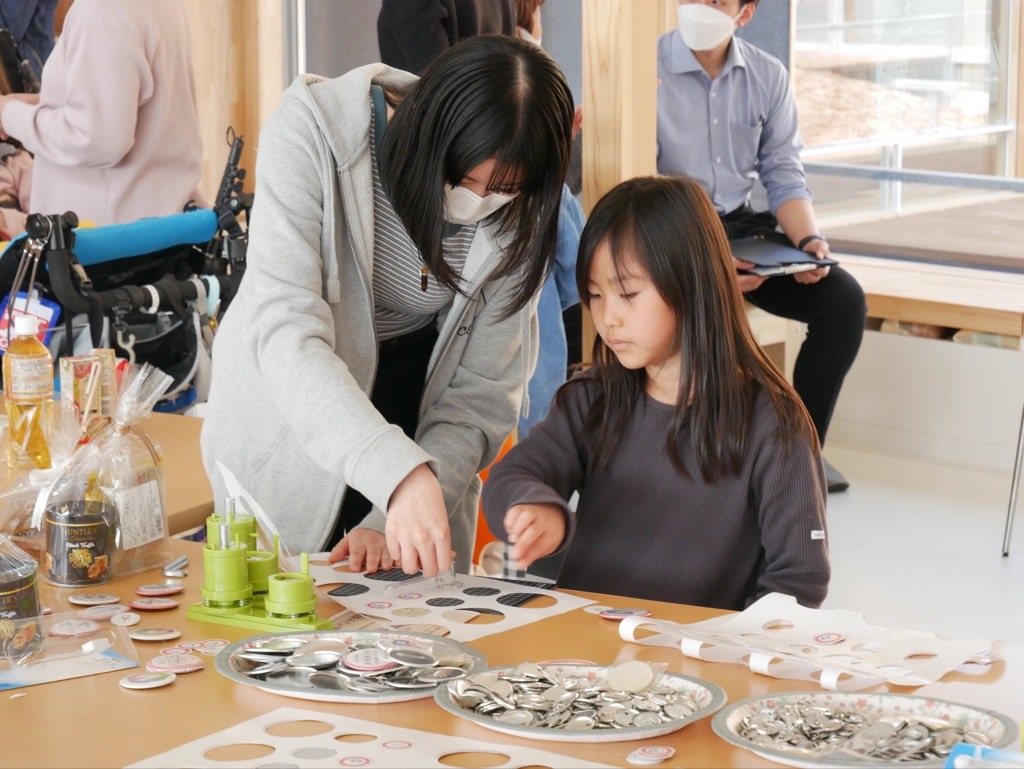 机に並べた材料を前に、大人と子どもが缶バッジを作っている